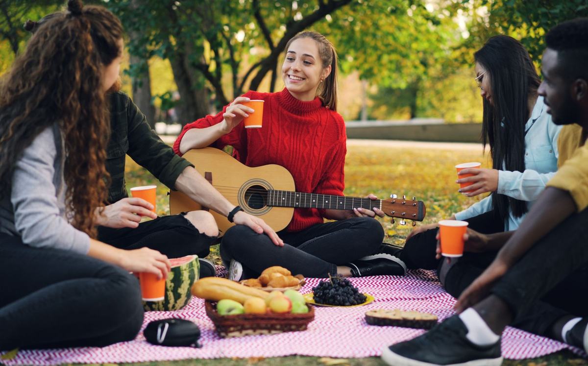 Friends sitting on a blanket in a park, one of them playing a guitar.